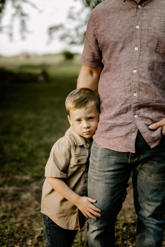 Boy holds his dad's leg and looks at the camera timidly.