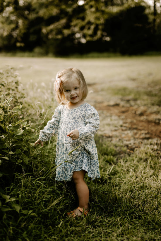 Toddler in a flowery dress stands in a field smiling at the camera for family photos.