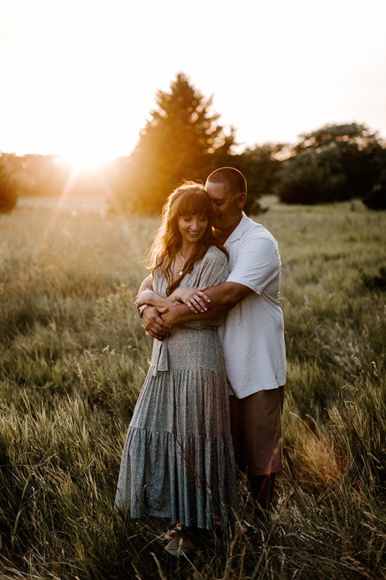 Husband embraces his wife during a family photo session. They are standing in a field with the sun shining in the background.