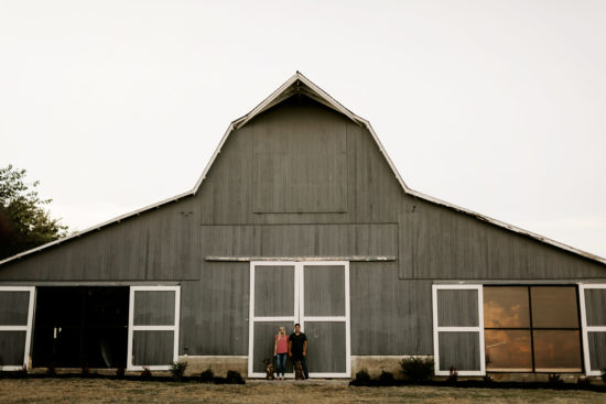 Couple holds hands in front of a barn with their two dogs at their side for their Kansas engagements photos