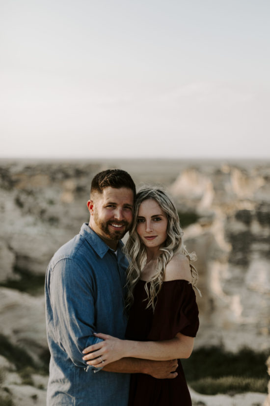 Couple embrace and smile gently, looking at the camera. The Little Jerusalem Badlands are in the background.