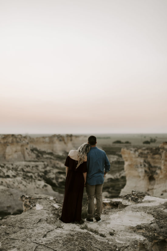A woman rests her head on her fiancé's shoulder, both are overlooking the Little Jerusalem badlands during their Kansas engagement photos