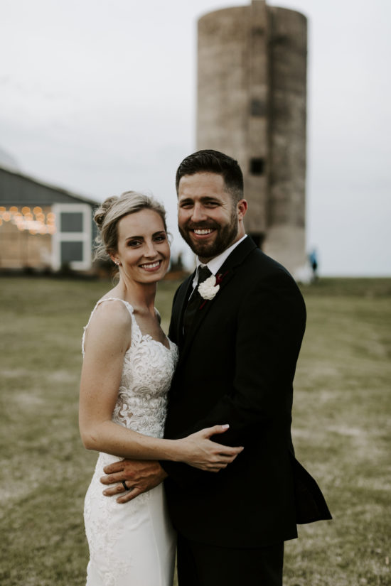 Bride and groom smile and hold each other in front of their Kansas farm barn and silo