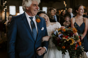 Bride laughs as she rests her head on her father's shoulder as he walks her down the aisle at her Jake and Abe's wedding