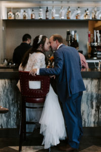 Bride and groom kiss while sitting a bar after their Abe and Jake's wedding