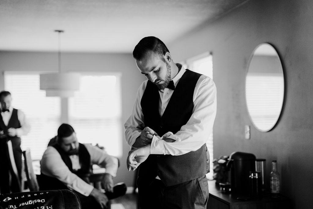 The groom stands in his living room, looking down while buttoning his shirt before before his Topeka KS wedding