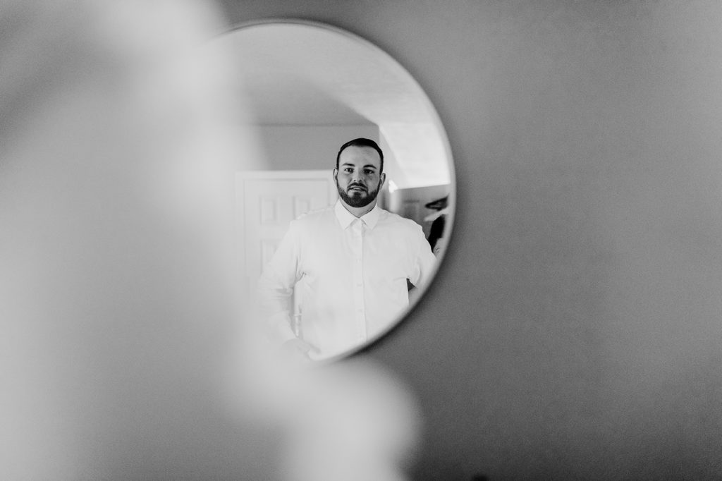 The groom looks in the mirror in his living room as he dresses for his wedding day