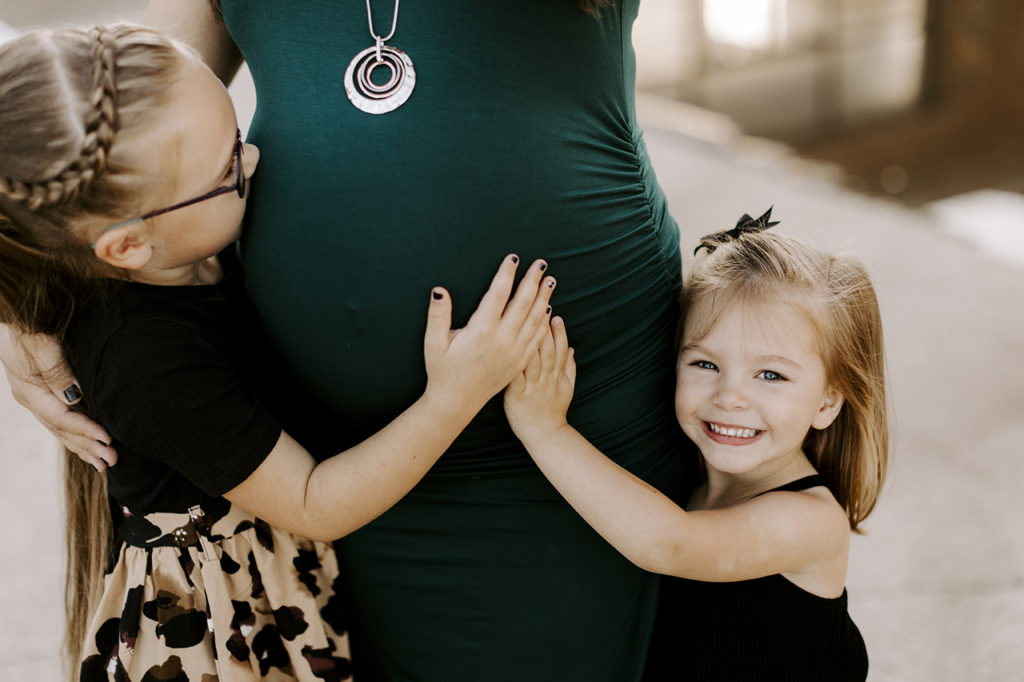 A girl holds her pregnant mom's belly and kisses it, while her sister holds mom's belly with a big grin on her face.