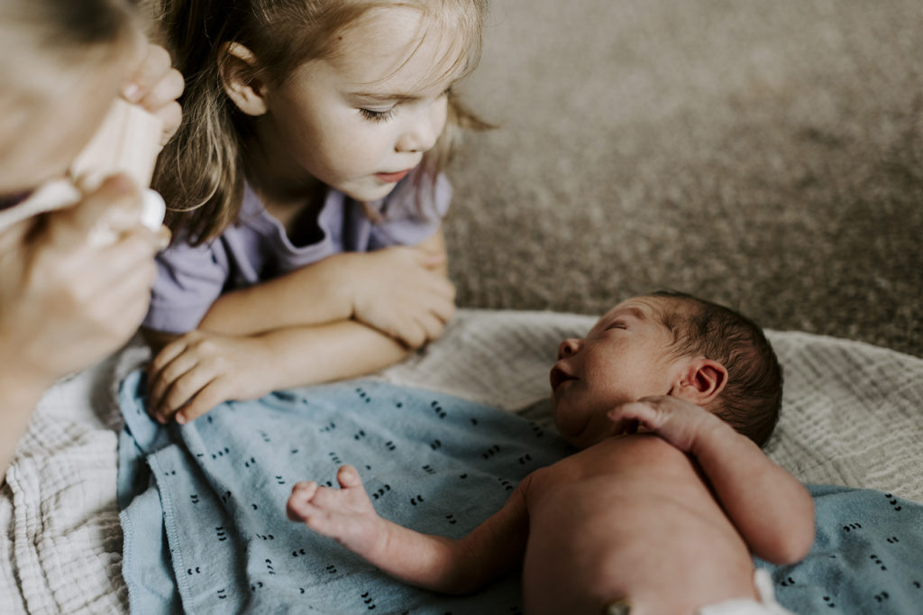 A newborn baby lays on blankets and looks up at his sisters, who are watching his every move.