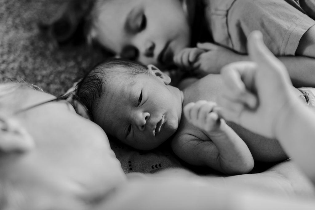 A newborn baby lays on blankets during his newborn session with his sisters snuggled up by him.