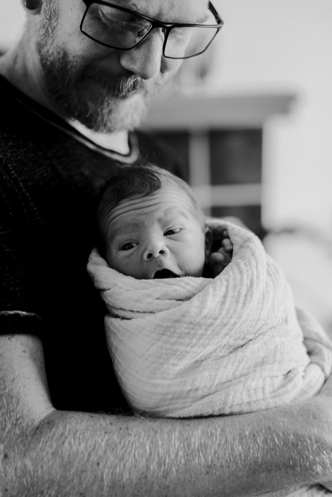 Dad holds his newborn, who is yawning and snuggled up in a wrap.