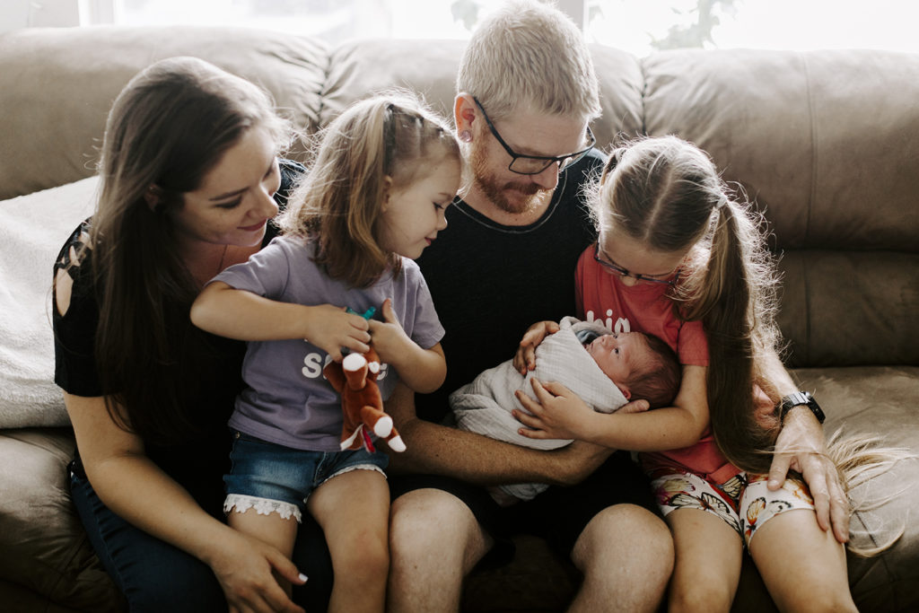 A dad holds his newborn son while surrounded by his family. The oldest daughter has her arms wrapped around baby.