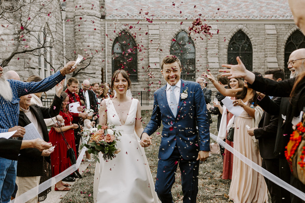 A bride and groom hold hands and smile while rose pedals are tossed in the air during their upscale Kansas City wedding.
