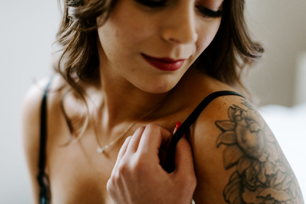 A woman smiles and holds the strap of her bra during her boudoir shoot in Lawrence KS