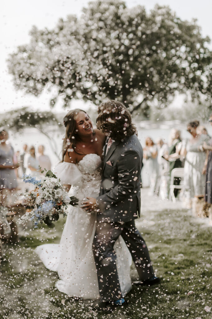 The bride and groom hold each other and laugh as confetti surrounds them during their Kansas lake house wedding.