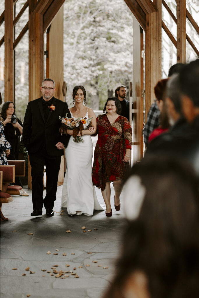 The bride is escorted down the aisle by her parents inside Marjorie Powell Allen Chapel at her Powell Gardens wedding.