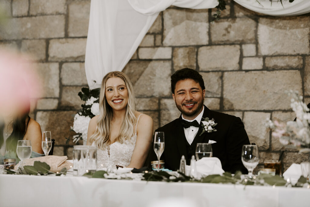 The couple smile and laugh as the father of the bride shares a toast for the happy couple